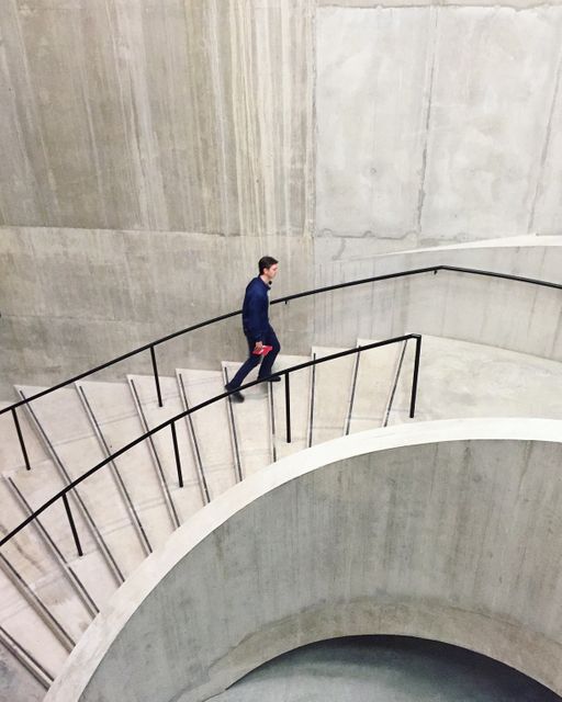 Man walking up a winding concrete staircase in a minimalist, modern building. The sleek design and neutral colors emphasize the architectural elements. Ideal for concepts related to modern architecture, interior design, minimalism, professional life, and urban environments. Useful for business presentations, architectural magazines, lifestyle blogs, and promotional materials highlighting contemporary spaces.
