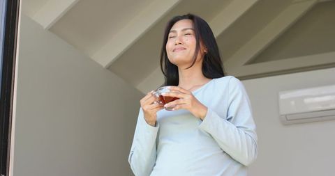Asian woman standing at doorway holding cup of tea enjoying calm morning light