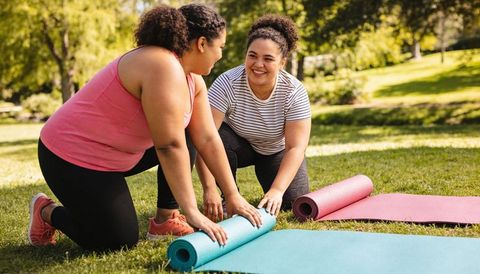 Rolling out yoga mats in sunny park, two women smiling and preparing outdoor workout