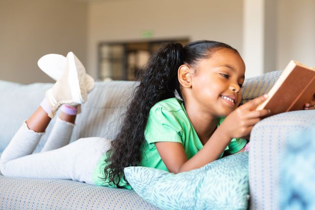 Young African American girl lying on sofa, smiling while reading a book. Ideal for educational materials, children's books, school promotions, and articles on childhood learning and relaxation.