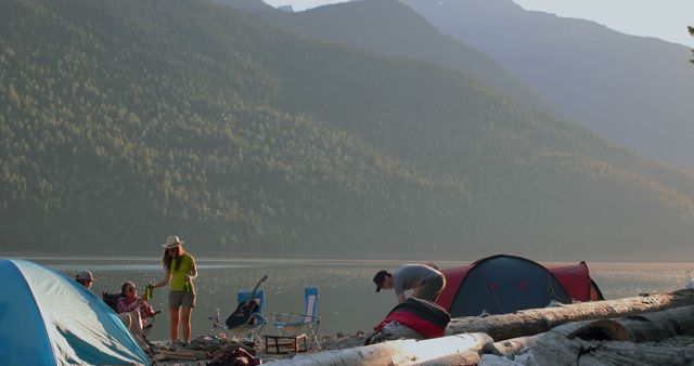 Group of friends enjoying a camping trip by a beautiful mountain lake during sunset. Camping tents set up along the lakeshore with logs and chairs around a natural campfire. Ideal for depicting friendship, outdoor adventure, and peaceful nature escapes. Useful for travel magazines, outdoor adventure promotions, nature-related content, and social media posts celebrating adventure and relaxation in nature.