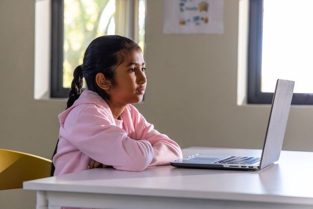 Asian schoolgirl studying with laptop at classroom desk wearing pink hoodie, e-learning