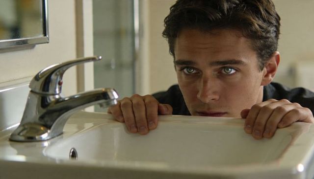 Teenager peering over sink gripping basin edge reflective introspective gaze in bathroom