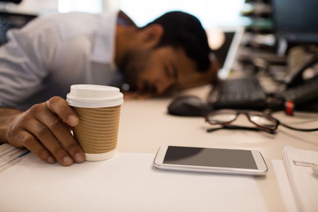 Businessman sleeping while holding disposable coffee up on desk at office
