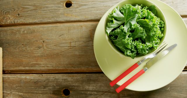 This colorful arrangement of a fresh green kale salad in a bowl is set on a rustic wooden table, complemented by a yellow plate and red-handled cutlery beside it. Ideal for promoting healthy eating, vegetarian recipes, organic lifestyles, food blogs, and restaurant menus.