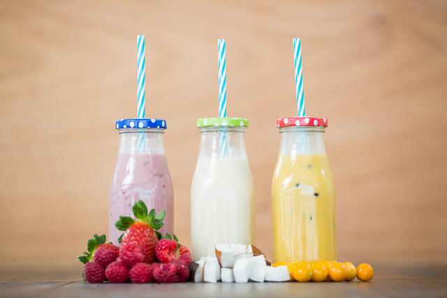 This image showcases three bottles of fruit smoothies with colorful lids and straws, accompanied by fresh chopped fruits including raspberries, coconut, and yellow berries, placed on a wooden board. Ideal for promoting healthy eating, diet plans, refreshing beverages, or smoothie recipes.