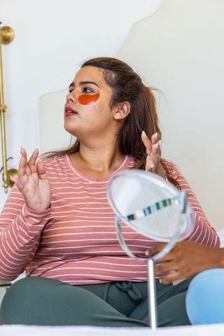 African american women sharing skincare routine applying orange under-eye patches on bed