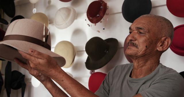 Happy senior bald biracial man with moustache holding hat in showroom at hat shop. Millinery, hats, local business, retail, shop, work, tradition and craft, unaltered.