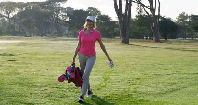 A woman golfer wearing a pink polo shirt and beige pants is walking on a green golf course carrying a set of golf clubs. The background features green trees and a bright, sunny sky. Perfect for use in articles and promotions related to sports, recreational activities, outdoor leisure, women's golfing, and healthy lifestyles.