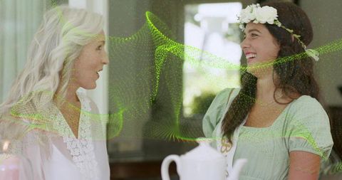 Senior mother and daughter sharing tea and smiles wearing floral crown with green particles