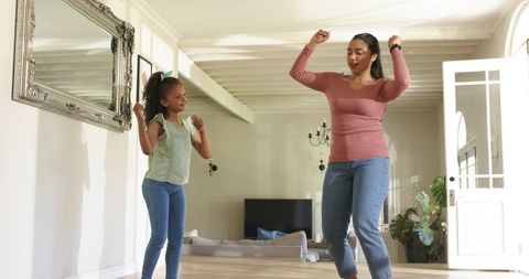 Mother and daughter dancing in sunlit living room on hardwood floor, casual home joy