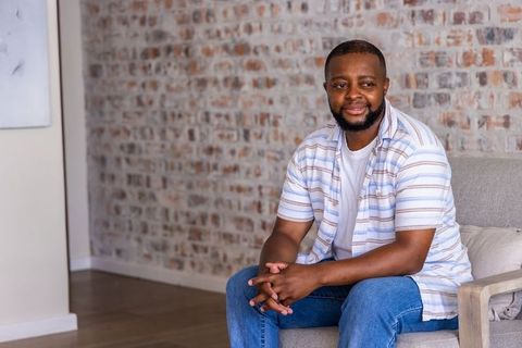 African man sitting in living room wearing striped shirt and jeans with exposed brick wall