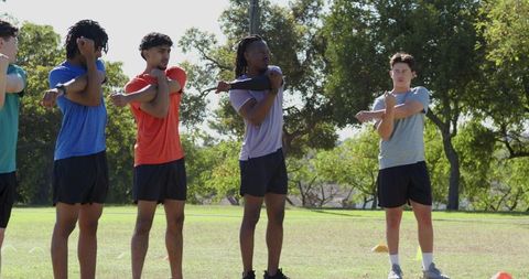 Diverse male teammates stretching shoulders during warm-up on sunny outdoor sports field