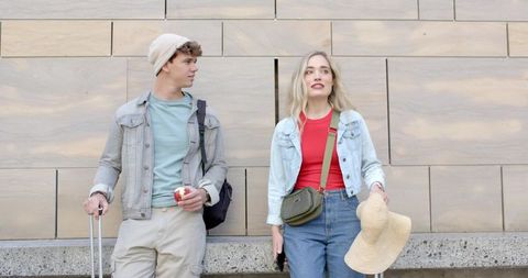 Young couple standing with luggage, denim jackets and summer accessories in urban plaza