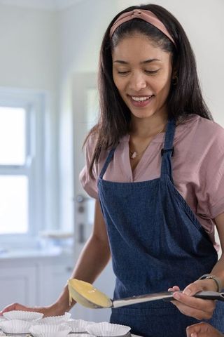 African american woman pouring cupcake batter in sunlit home kitchen wearing denim apron