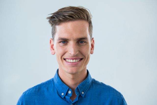 This image features a young man smiling confidently while wearing a blue shirt against a white background. Ideal for use in professional profiles, social media, advertisements, and websites promoting positive and friendly environments. It can also be used in articles or blogs about fashion, lifestyle, and personal development.