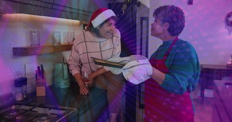 Holiday baking laughing woman in santa hat sitting on counter while friend removing tray