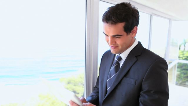 Young businessman in a suit using a tablet while standing near large windows with a seaside view. Ideal for depicting modern technology in business settings, corporate life, and professional work environments.