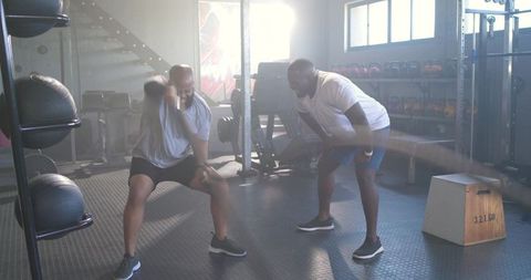 Two men performing kettlebell swings while trainer coaching in sunlit gym