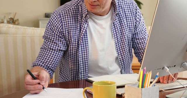 Man in casual attire working from home office, sitting at desk with computer and documents. This scene can be used in articles and posts about remote work, productivity, entrepreneurship, home office setups, and work-life balance. Ideal for illustrating blog posts, websites, and presentations related to business and working remotely.