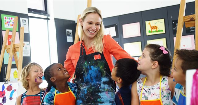 Art teacher interacting with a group of smiling children in a classroom filled with paintings. Could be used for educational content, promotional material for schools or art programs, or articles on creative teaching methods and inclusivity in education.