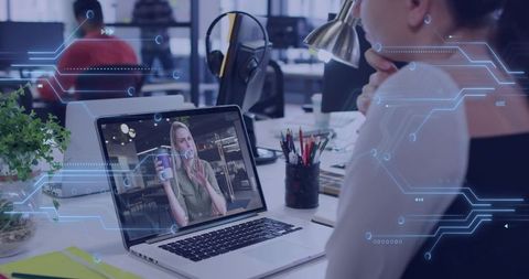 Professional watching remote video call on laptop in open-plan office with tech overlay