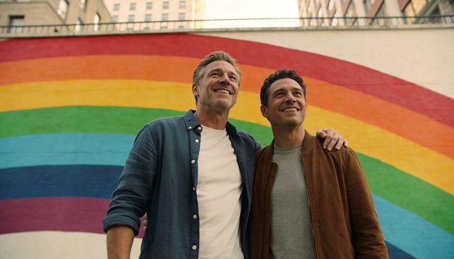 Smiling gay couple embracing before vibrant rainbow mural celebrating pride and urban love