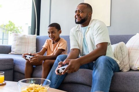 African american dad and son playing video games on couch with snacks and playful rivalry