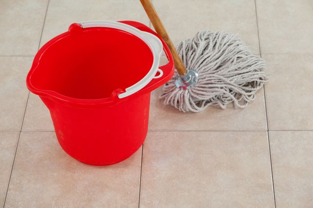 This image shows an empty red bucket and a mop on a tile floor. It is ideal for illustrating concepts related to household chores, cleaning routines, and sanitation. It can be used in articles, blogs, or advertisements about home maintenance, janitorial services, and cleanliness.