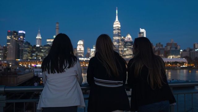Three friends leaning on waterfront railing watching new york skyline at dusk