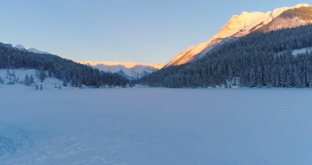 A serene winter landscape showcases a frozen lake surrounded by snow-covered trees and mountains bathed in the warm glow of sunrise. The tranquil setting evokes a sense of peace and the beauty of nature in its winter attire.