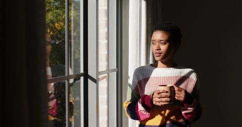 Woman in colorful sweater holding mug and gazing through window