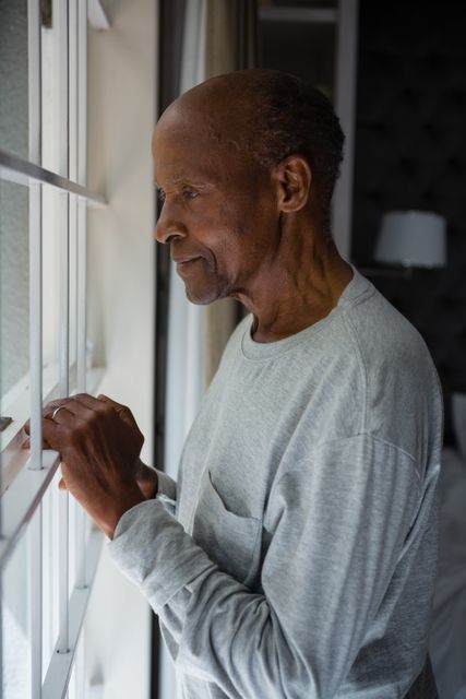 This image captures a senior man standing by a window, looking outside with a thoughtful expression. Ideal for use in articles or advertisements related to aging, retirement, senior living, mental health, and lifestyle. It conveys themes of contemplation, solitude, and peacefulness.