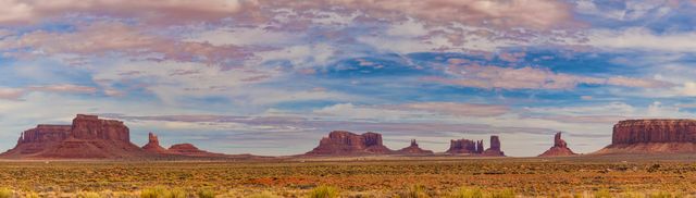 Panoramic view showcasing the vast and iconic landscape of Monument Valley, featuring unique red rock formations under a dramatic, partly cloudy sky. Ideal for travel promotions, landscape photography collections, and backgrounds for presentations or websites reflecting natural beauty and adventure.