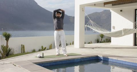 Indian man stretching on yoga mat by poolside terrace with hammock and mountain lake view