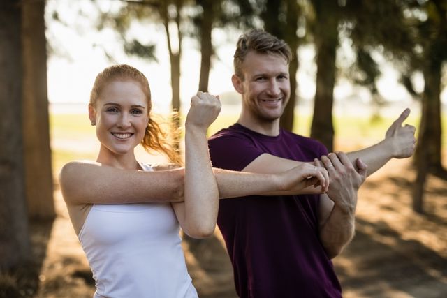 Couple stretching arms and smiling in outdoor setting with trees and sunlight. Ideal for promoting fitness, healthy lifestyle, outdoor activities, and wellness programs. Can be used in advertisements, blogs, and social media posts related to exercise and nature.