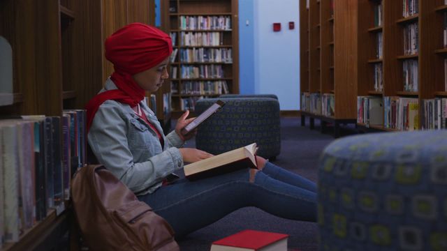 Young Asian female student in red hijab studying in a library while sitting on the floor, reading a book and using her smartphone. She is wearing casual clothing, including a denim jacket. This scene is ideal for use in content related to education, studying, online learning, young women's empowerment, and the importance of libraries.