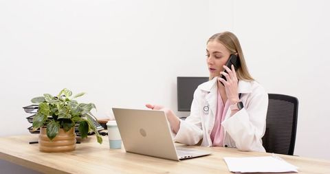Female doctor talking on smartphone while gesturing at laptop in modern clinic office