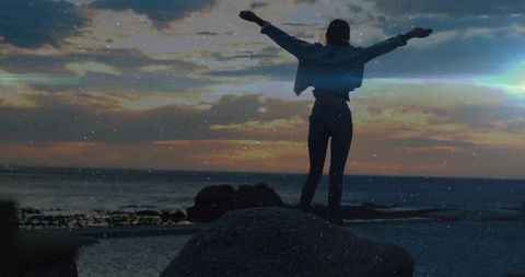 Woman standing on boulder raising arms at sunset silhouette over tidal pools and horizon
