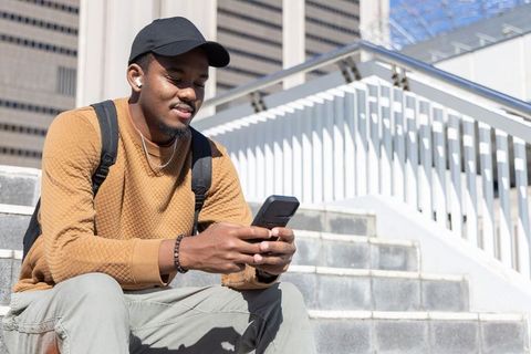 African american man sitting on city steps using smartphone wearing backpack and cap