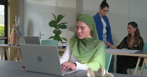 Focused woman wearing green hijab working on laptop in modern coworking collaborative space