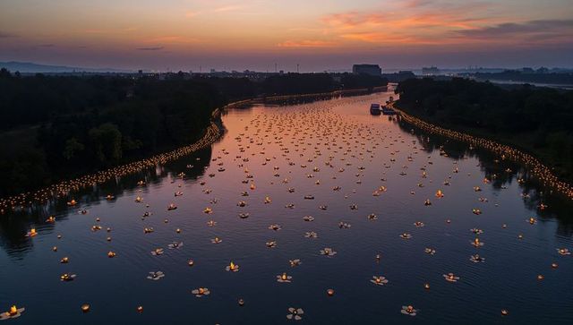 Aerial dusk panorama of drifting lanterns filling river with glowing reflections and shore lights