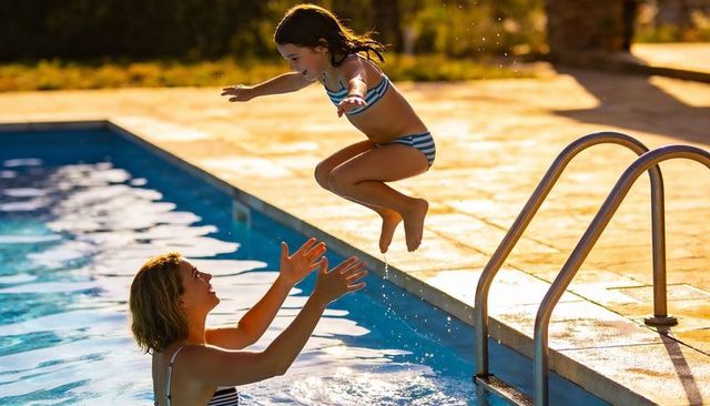 Parent catching daughter jumping into backyard pool at golden hour, striped swimsuit fun