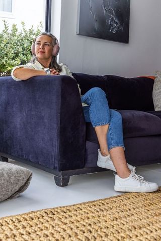Middle-aged woman relaxing on navy velvet sofa wearing headphones and gazing out window