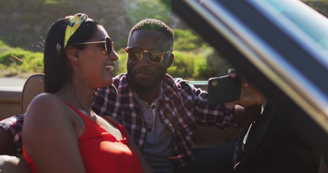 Couple enjoying a road trip in a convertible car. Both are wearing sunglasses and smiling, with the woman in a red top and man in a checked shirt. They are taking a selfie while surrounded by an outdoor, natural background. Perfect for use in themes of travel, adventure, relationships, leisure, and summer holidays.