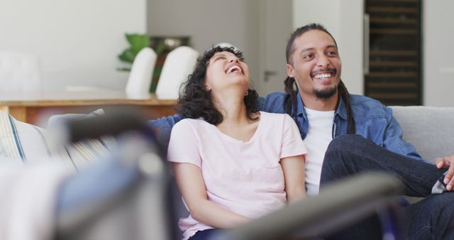 Happy biracial couple in living room watching tv and laughing, with her wheelchair in foreground. wellbeing and domestic lifestyle with physical disability.
