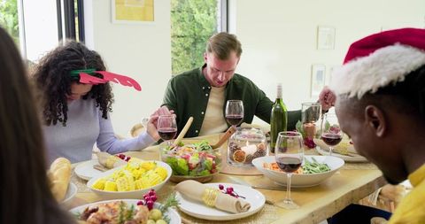 Diverse friends holding hands and bowing heads around sunlit holiday meal sharing gratitude