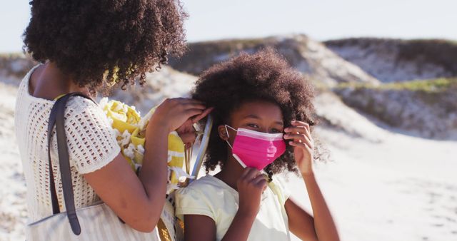 Mother adjusts her daughter's face mask while at the beach. They are both enjoying a sunny day outdoors. The image can be useful for promoting family safety during outdoor activities, health and hygiene products, and pandemic-related awareness campaigns.