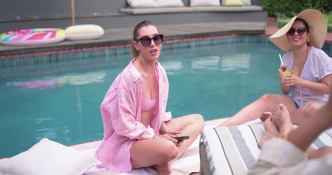 Diverse female friends relaxing poolside in swimwear with cocktails and smartphone