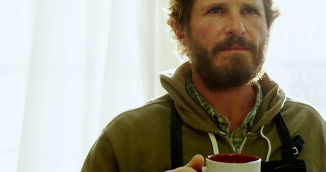A middle-aged Caucasian man with a beard is holding a red mug, with copy space. He appears contemplative as he gazes out of the frame, enjoying a quiet moment.